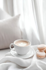 Fototapeta premium Cup of cappuccino with latte art and pastel macarons on a white plate, placed on a soft fabric near a pillow. Cozy and minimalistic breakfast scene with natural light and airy curtains
