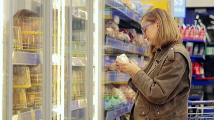 Focused woman choosing frozen food in supermarket aisle, reading labels for healthy options. Cold display offers variety for informed decisions
