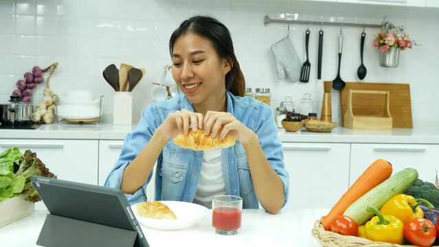 Happy asian woman eating healthy food drinks watch online movie. Women enjoy video call web conference live streaming home kitchen use digital tablet. Female use smart phone online call social media