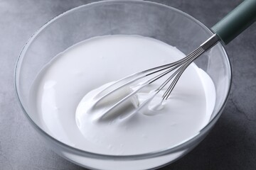 Whisk and whipped cream in bowl on grey table, closeup