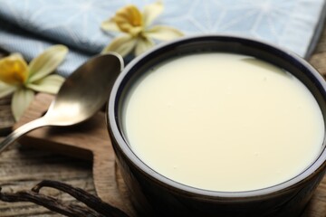Tasty condensed milk, spoon, vanilla pods and flowers on wooden table, closeup