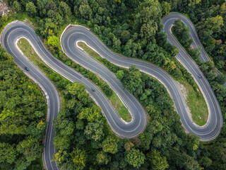 zenithal aerial view of a winding road in a green forest as cars pass by on the road in Romania.