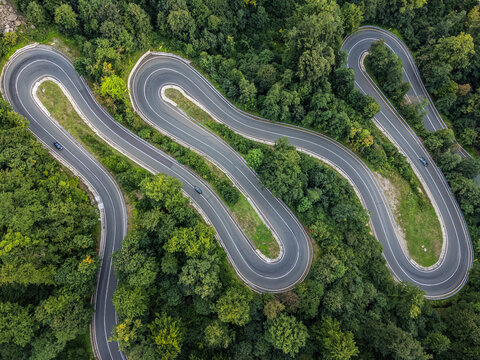 zenithal aerial view of a winding road in a green forest as cars pass by on the road in Romania.