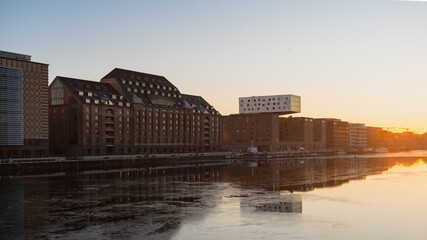 Sunset view of the Spree River in Berlin with historic brick warehouse buildings and a modern, cantilevered hotel, reflecting in the calm water