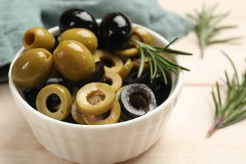 Different delicious marinated olive rings, whole ones and rosemary in bowl on light wooden table, closeup