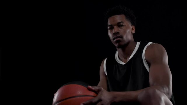 Basketball player sitting on a bench. Looking at camera, Concentrated african american sports man against black background.