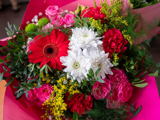A vibrant bouquet of mixed flowers, including red gerberas, white chrysanthemums, pink and red carnations and delicate yellow flowers, wrapped in pink paper.