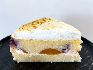 A slice of baked rice pudding with apricots and blueberries sits on a black textured plate on a wooden table. The top has a golden-brown caramelized layer with small droplets.