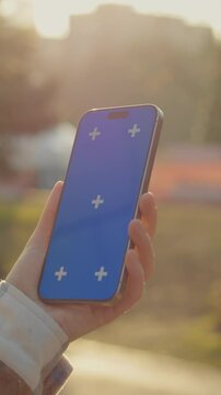 Vertical Screen: Young woman is holding a smartphone with a blue chroma key screen outdoors in a sunny day