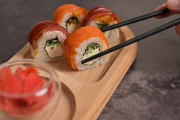 Set of sushi roll with rice and fish on wooden serving board. Chopsticks holding sushi roll. Close up. Dark black texture background with copy space. Japan menu. Asian food