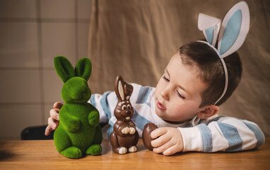 Boy with bunny ears 5 years old plays with Easter bunnies at table: with chocolate bunny and green...