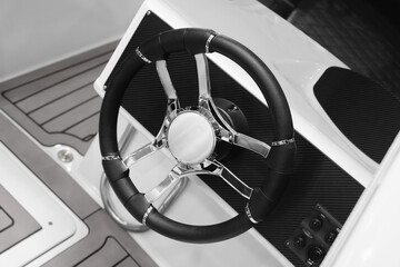 A black steering wheel with a silver rim. The wheel is on a boat. The boat is white