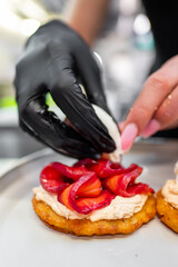 A chef in black gloves expertly garnishes a golden fritter topped with creamy spread and vibrant red peppers on a stainless steel tray.