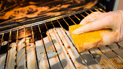 Cleaning the oven from heavy fat in the kitchen. Hand in a household glove and with a yellow sponge for cleaning, out of focus on the background of the oven. Anti-grease for cooking surfaces