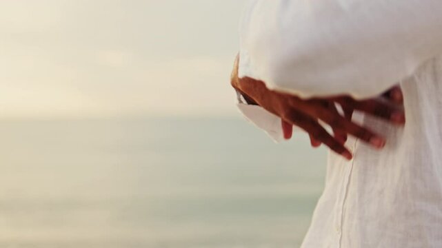 A man in a white shirt practices Tai Chi-like hand movements by the sea at sunset. Meditative practice, slow motion
