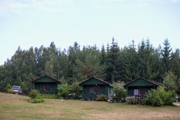 
three small wooden houses with dark green roofs and walls, located in the forest with a small car on the left.