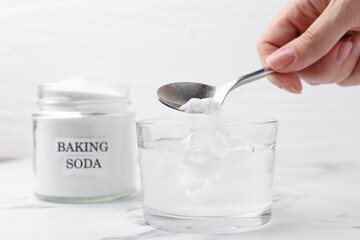 Woman adding baking soda to glass of water at white marble table, closeup