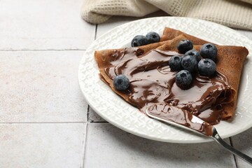 Delicious chocolate crepes with sweet paste and blueberries on light tiled table, closeup. Space for text