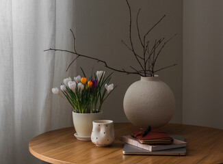 Cozy spring home still life - crocuses in a pot, a round ceramic vase, a laptop, a cup of coffee on a round wooden table in the living room