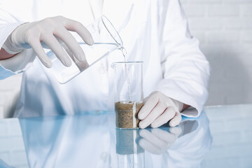 Laboratory testing. Scientist pouring liquid into beaker with soil sample at table indoors, closeup