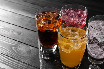 Refreshing soda water of different flavors in glasses with ice cubes on black wooden table, space...