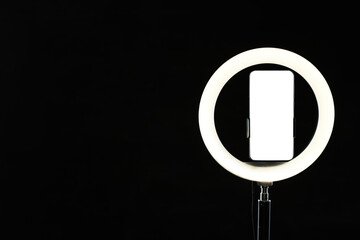 Ring lamp with smartphone on black background, space for text