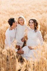 A pregnant woman and a man are hugged by their daughter, in a field for growing wheat - the concept...