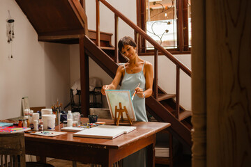 A female artist in a long blue dress draws a picture near a wooden table with materials for creativity. Concept of teaching drawing and hobbies