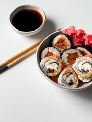 Sushi rolls in a bowl with pink ginger and soy sauce and wooden sticks on white table