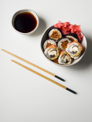Sushi rolls in a bowl with pink ginger and soy sauce and wooden sticks on white table