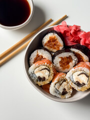 Sushi rolls in a bowl with pink ginger and soy sauce and wooden sticks on white table close up