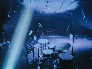 A dimly lit stage featuring a drum kit, microphones, and audio equipment under moody blue lighting....