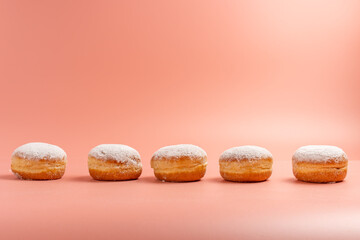 A row of powdered donuts is lined up on a pink background. The donuts are all the same size and shape, and they are all covered in powdered sugar. The background is a light pink color