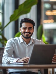 Smiling indian businessman working on laptop in modern office lobby space. Young indian student using computer remote studying, watching online webinar, zoom virtual training on video call meeting.