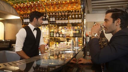 Focusing smiling smart bartender serving gin fizz or tonic soda at luxurious bar, engaging lively conversation with VIP customer couple at back view in atmosphere at lighting warm nightclub. Vinosity.