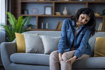 A young woman clutches her knee in pain while sitting on the couch in her living room.