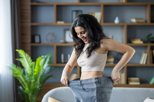 A young woman happily shows her oversized jeans, celebrating her successful weight loss journey.