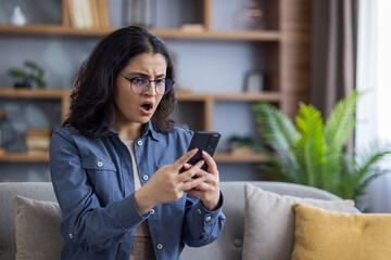 A young woman gasps in shock, staring intently at the screen of her smartphone while relaxing on...
