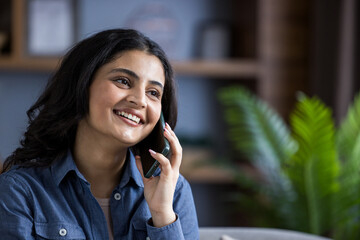 A happy woman smiles during a lively phone conversation indoors, wearing a casual denim shirt.
