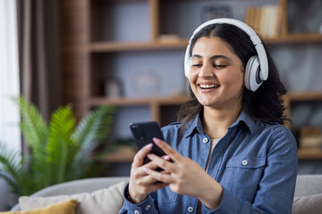 A cheerful woman enjoys music on her smartphone with headphones in a cozy living room.