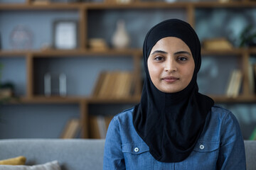 Portrait of a young woman wearing a hijab, seated in her modern living room with a serene...