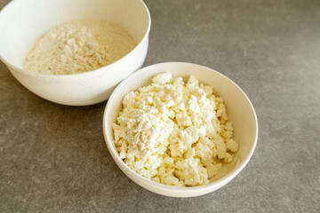 Two white bowls filled with ingredients for baking, one containing fresh cottage cheese and the other filled with flour. 