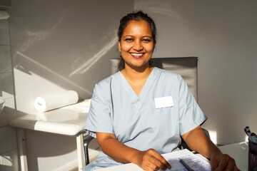 Portrait of smiling female healthcare worker wearing medical scrubs sitting near table in clinic