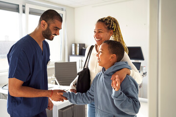 Happy male healthcare worker shaking hands of boy leaving medical examination room with mother at hospital