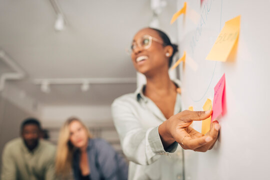 Smiling young businesswoman holding adhesive note on whiteboard during discussion in meeting room at office