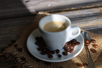 A cup of coffee and coffee beans on the saucer, placed on a burlap sack on an old wooden table. 