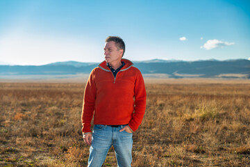 Rugged man rancher in orange pullover fleece standing in open grassland with Rocky Mountains in background