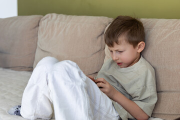 Fototapeta premium Young boy focused on smartphone screen. A young boy sits on a beige couch, intently looking at his smartphone, deeply engaged in a game or digital content at home.