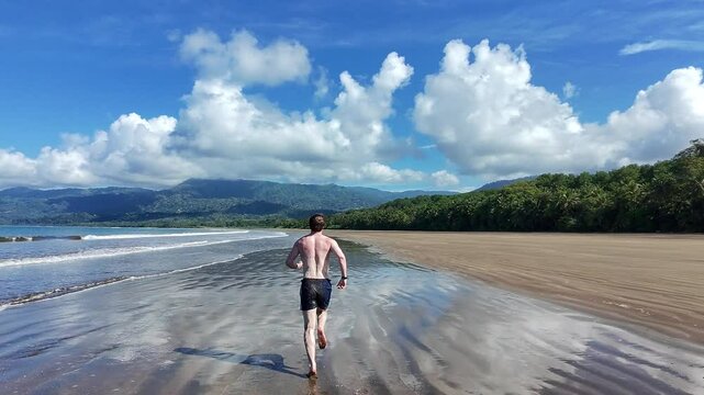 Athletic man running along Costa Rica shore line beach - camera follows behind man
