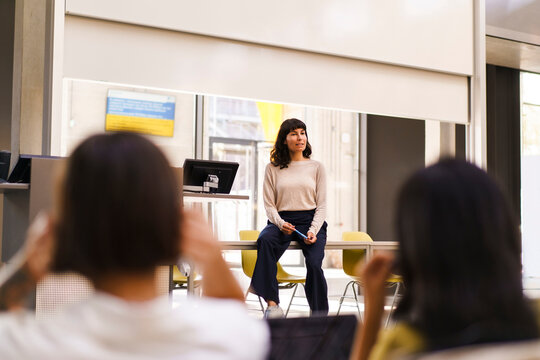 Female students listening to teacher sitting on table while explaining in lecture hall at college